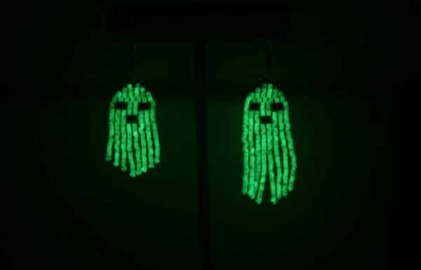 Earrings hanging on a black display in the dark and glowing. There is a single dangle of each of the Spooky Ghost lengths hanging from a black display.