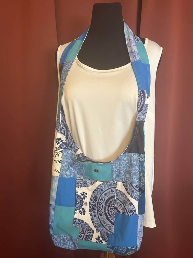 Lining view - Shades of blue cotton fabric in patchwork design. Pocket on each side 6" x 6" perfect for cell phone, sunglasses or keys. A 2" wide strap for easy crossbody carry. A magnetic snap secures belongings.