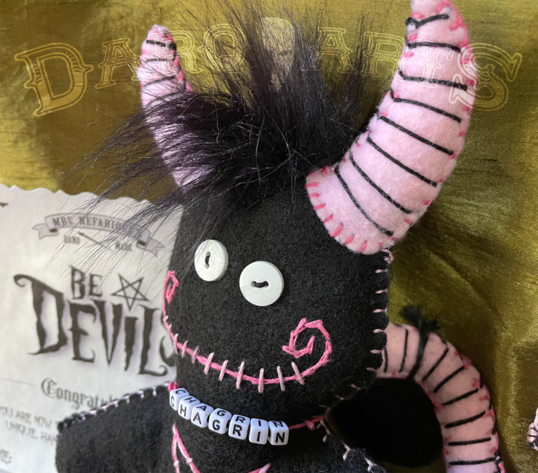Bedevils - Chagrin. Black soft felt body with piggy pink horns, tail, hooves and reversed pentagram. Close up of face.