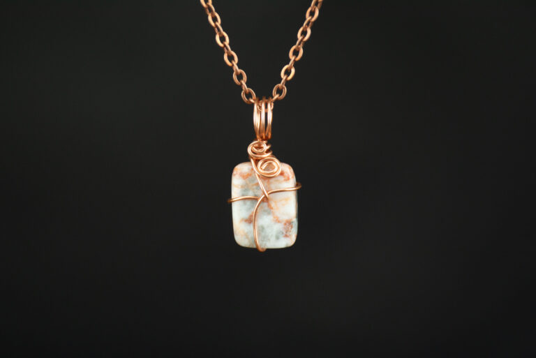 Mottled orange and white jasper stone wrapped minimally in copper wire. This is hanging from a copper chain on a black background.