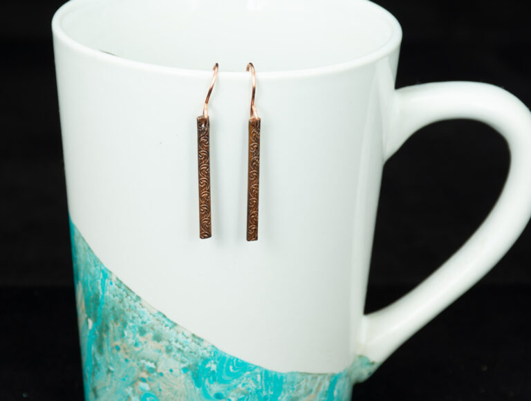 Thin bar stamped copper earrings hanging from the lip of a white coffee mug with an abstract blue design at an angle near the bottom.