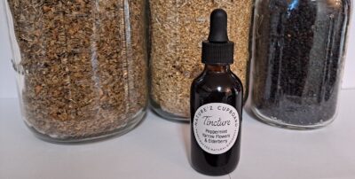 Amber dropper bottle labeled "Nature’z Cupboard Tincture Peppermint Yarrow Flowers and Elderberry" in front of glass jars filled with dried herbs.