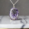 Chevron Amethyst pendant with a silver AA Recovery Symbol. The pendant is on a silver chain and it is shown hanging above a gray and white marble stone.