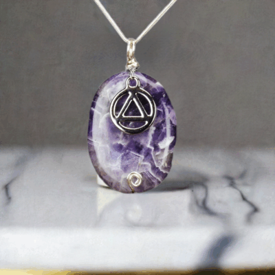 Chevron Amethyst pendant with a silver AA Recovery Symbol. The pendant is on a silver chain and it is shown hanging above a gray and white marble stone.