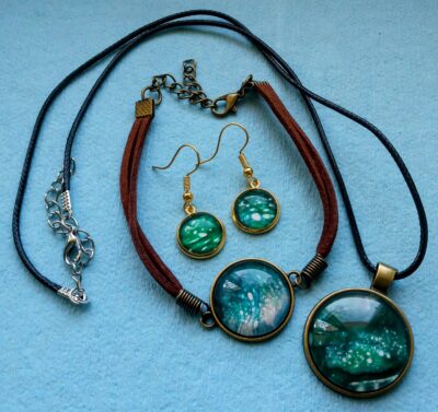 A beautiful set of fluid art jewelry: there is a necklace, bracelet, and pair of earrings in shades of green, teal, and white. The bezels are circular and gold.