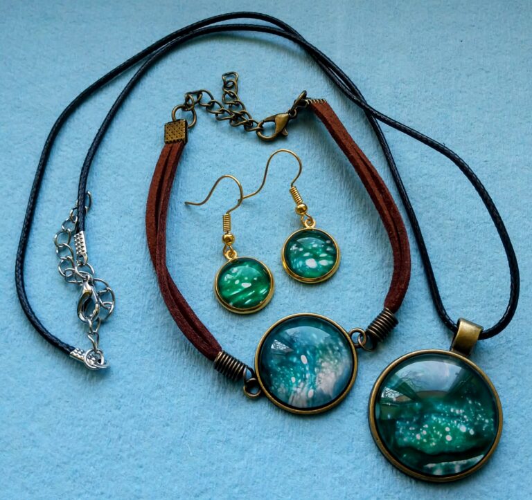 A beautiful set of fluid art jewelry: there is a necklace, bracelet, and pair of earrings in shades of green, teal, and white. The bezels are circular and gold.