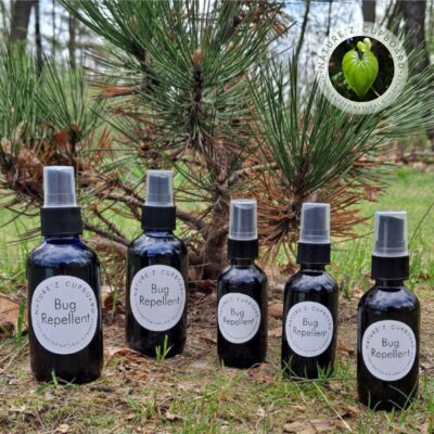 Five dark glass spray bottles labeled "Bug Repellent" from Nature’z Cupboard arranged in a row on forest ground in front of pine trees.