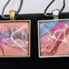 Two square paint dip pendants side by side: the one on the left has a bronze trays, and the one on the right is silver. The colors used in the paints are shades of pink, purple, blue, and red.