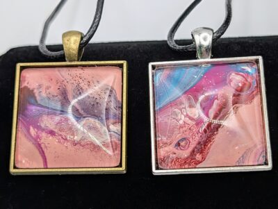 Two square paint dip pendants side by side: the one on the left has a bronze trays, and the one on the right is silver. The colors used in the paints are shades of pink, purple, blue, and red.