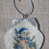 Handmade coastal snowman ornament on a scallop seashell with blue winter artwork and a light-blue ribbon hanger