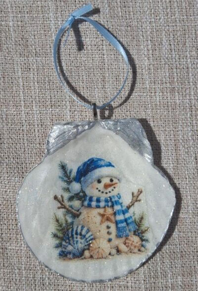 Handmade coastal snowman ornament on a scallop seashell with blue winter artwork and a light-blue ribbon hanger